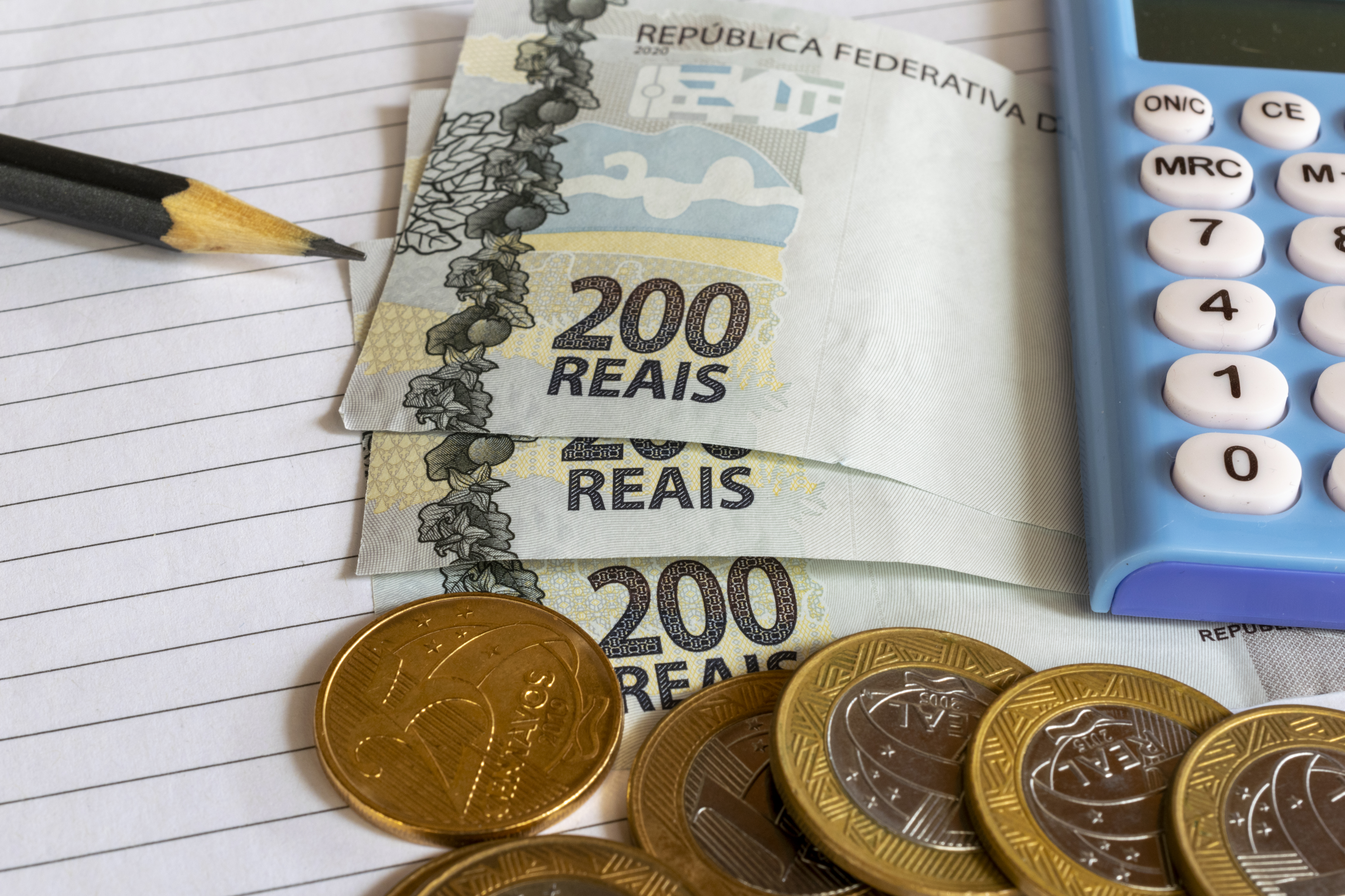 Notas de duzentos reais, moedas, lápis e calculadora sobre papel pautado, ilustrando o cálculo e a tributação de recursos financeiros.