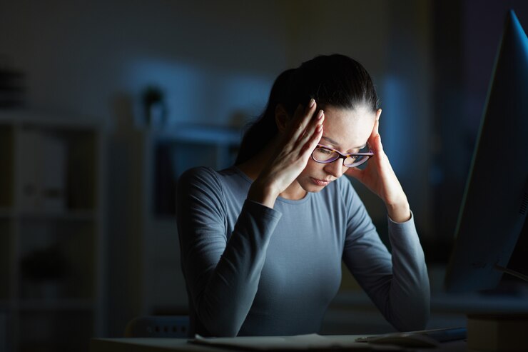 Mulher de óculos trabalhando em escritório mal iluminado à noite, segurando a cabeça com as mãos devido ao estresse e preocupação com a contabilidade e impostos.