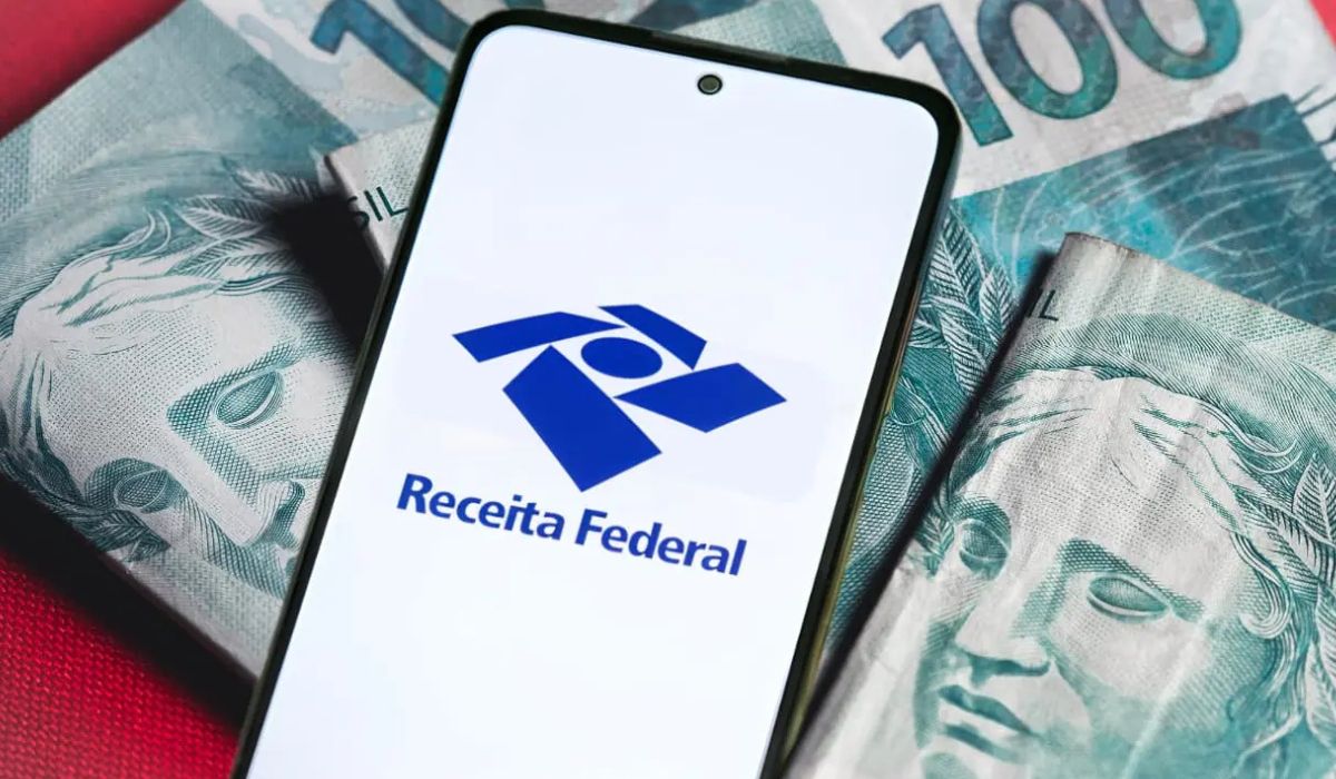 Celular exibindo o logotipo da Receita Federal sobre notas de dinheiro brasileiro, ilustrando a Restituição do Imposto de Renda.