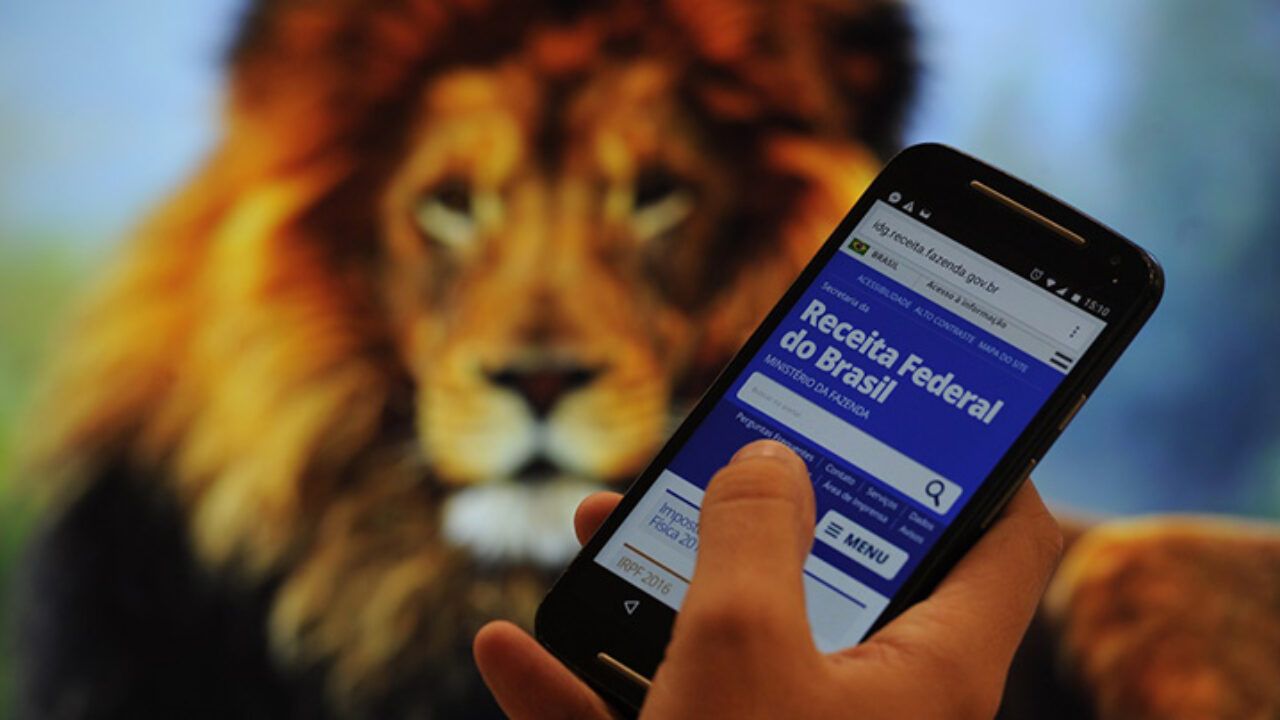 Pessoa acessando o site da Receita Federal pelo celular para consultar a restituição do Imposto de Renda, tendo ao fundo a imagem desfocada de um leão.