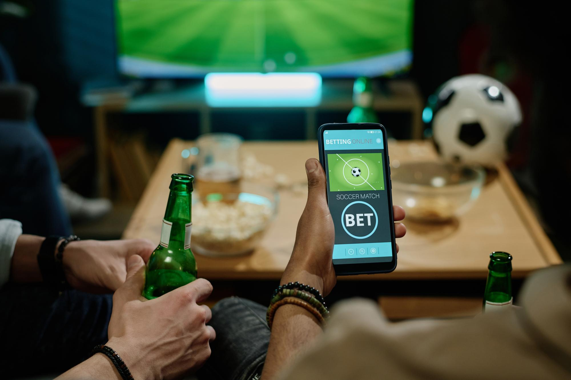 Pessoa segurando celular com aplicativo de apostas esportivas e garrafa de cerveja, enquanto assiste a jogo de futebol na TV com amigos.