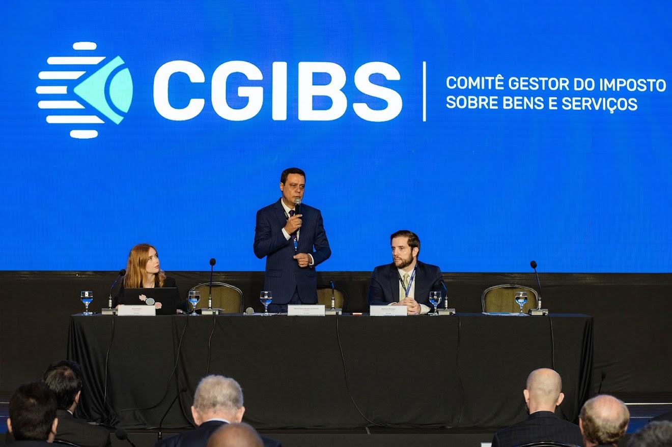 Mesa de autoridades discursando em evento oficial sobre a reforma tributária e a criação do CG | IBS, com o público assistindo de costas.