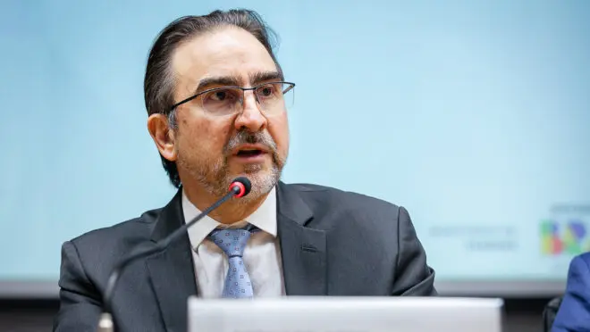 Retrato de um homem de óculos e terno falando ao microfone em um evento institucional sobre a reforma tributária.