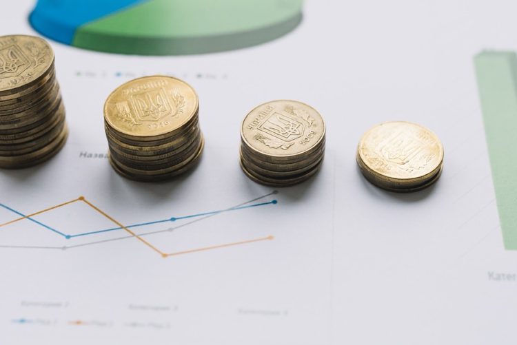 Pilhas de moedas douradas decrescentes sobre gráficos financeiros, ilustrando projeções econômicas e a reforma tributária em 2027 focada na cobrança por consumo.