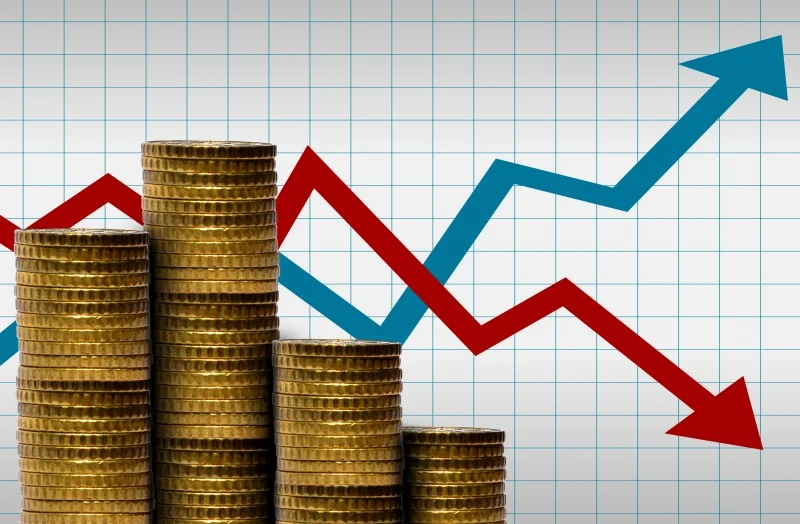 Pilhas de moedas douradas sobrepostas diante de um gráfico quadriculado com setas azul e vermelha indicando alta e baixa, ilustrando o impacto financeiro da taxa das blusinhas.