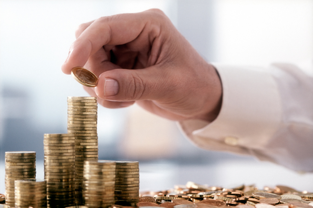 Mão adicionando uma moeda a uma pilha, simbolizando o controle e impacto fiscal de precatórios nas empresas.