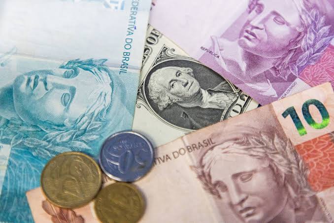 Cédulas de reais e notas de dólar misturadas com moedas sobre uma mesa, ilustrando o impacto da cotação da moeda americana no caixa da empresa.