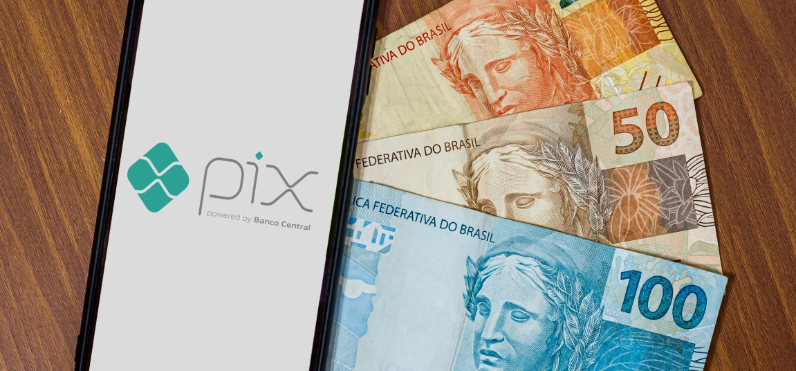 Smartphone exibindo a logo do Pix ao lado de notas de Real brasileiro sobre uma mesa de madeira.
