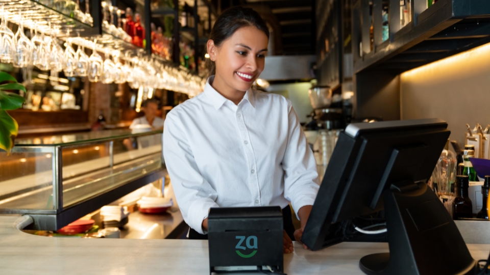 Atendente sorridente opera o caixa de um restaurante, simbolizando o desafio dos bares e restaurantes diante da Reforma Tributária.