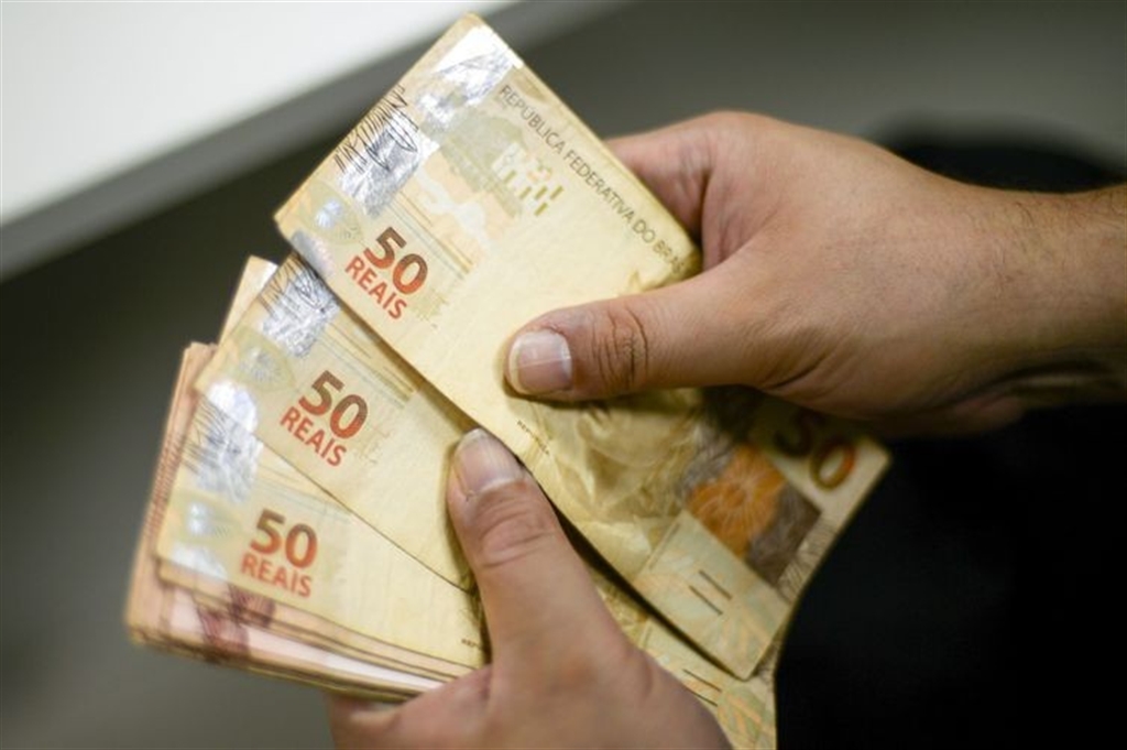 Mãos segurando um maço de notas de cinquenta reais, ilustrando o impacto financeiro das bets e das eleições 2026 no orçamento.