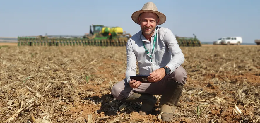 Produtor rural sorri agachado em lavoura com trator ao fundo, ilustrando os impactos da Reforma Tributária no agronegócio.