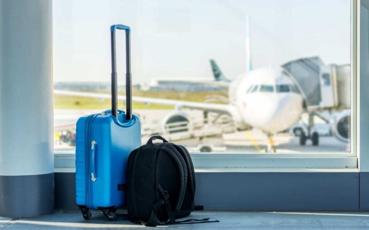 Mala azul de viagem e mochila preta no chão de um aeroporto, com um avião estacionado ao fundo na pista visto através de uma grande janela de vidro.
