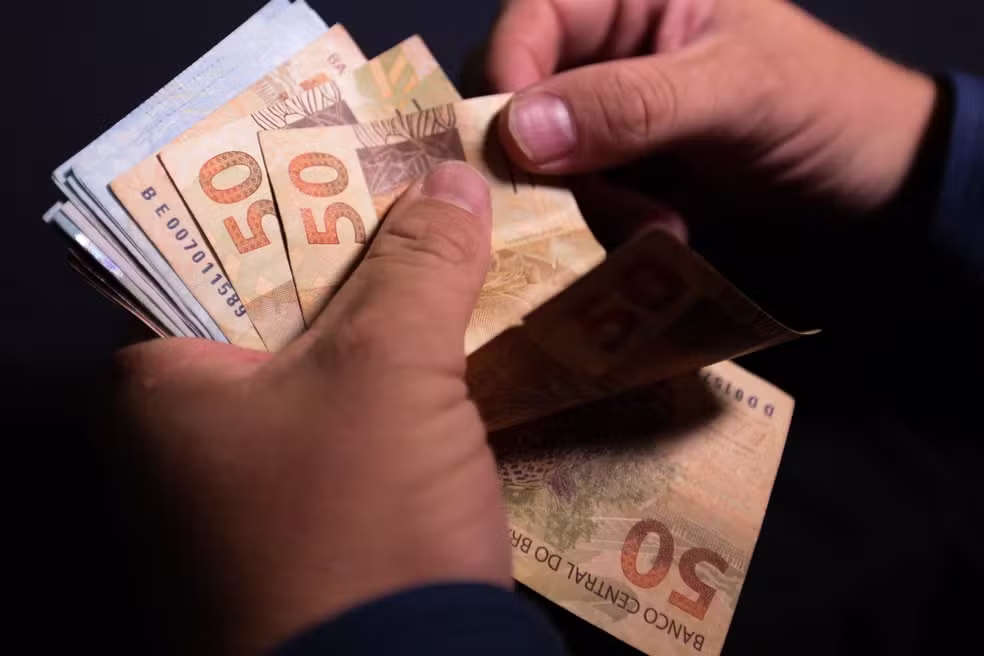 Mãos contando notas de dinheiro brasileiro, ilustrando restrições financeiras e a proibição da recuperação judicial para devedores contumazes.