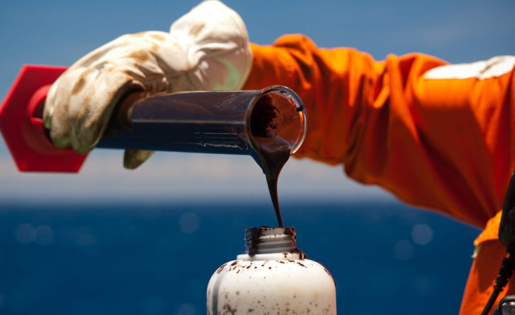 Trabalhador com uniforme laranja e luva de proteção despeja amostra de petróleo bruto em um frasco em alto-mar, em meio à discussão sobre o imposto de exportação de petróleo.