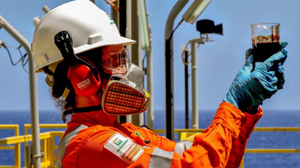 Trabalhador com EPI segura um recipiente com petróleo bruto em uma plataforma de exploração no mar.