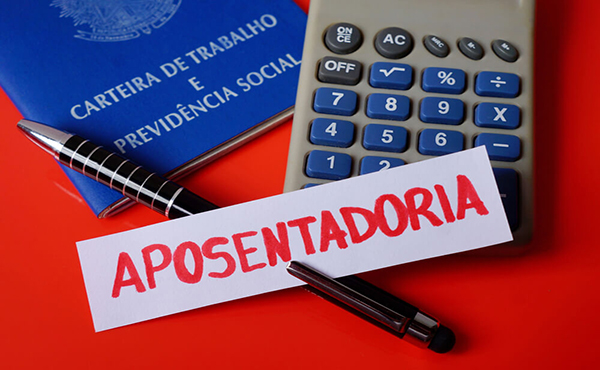 Carteira de trabalho azul, calculadora e uma caneta sobre fundo vermelho ao lado de um papel com a palavra aposentadoria, ilustrando o impacto das regras previdenciárias na empresa.