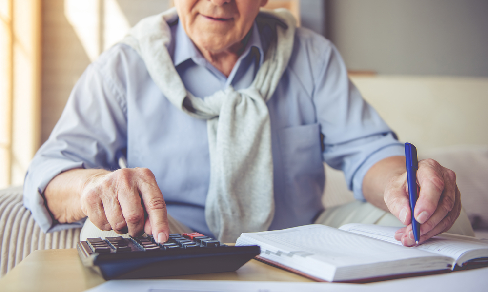 Homem idoso usa uma calculadora e anota em um caderno, ilustrando o planejamento financeiro para a aposentadoria com 67 anos e o impacto nas empresas.