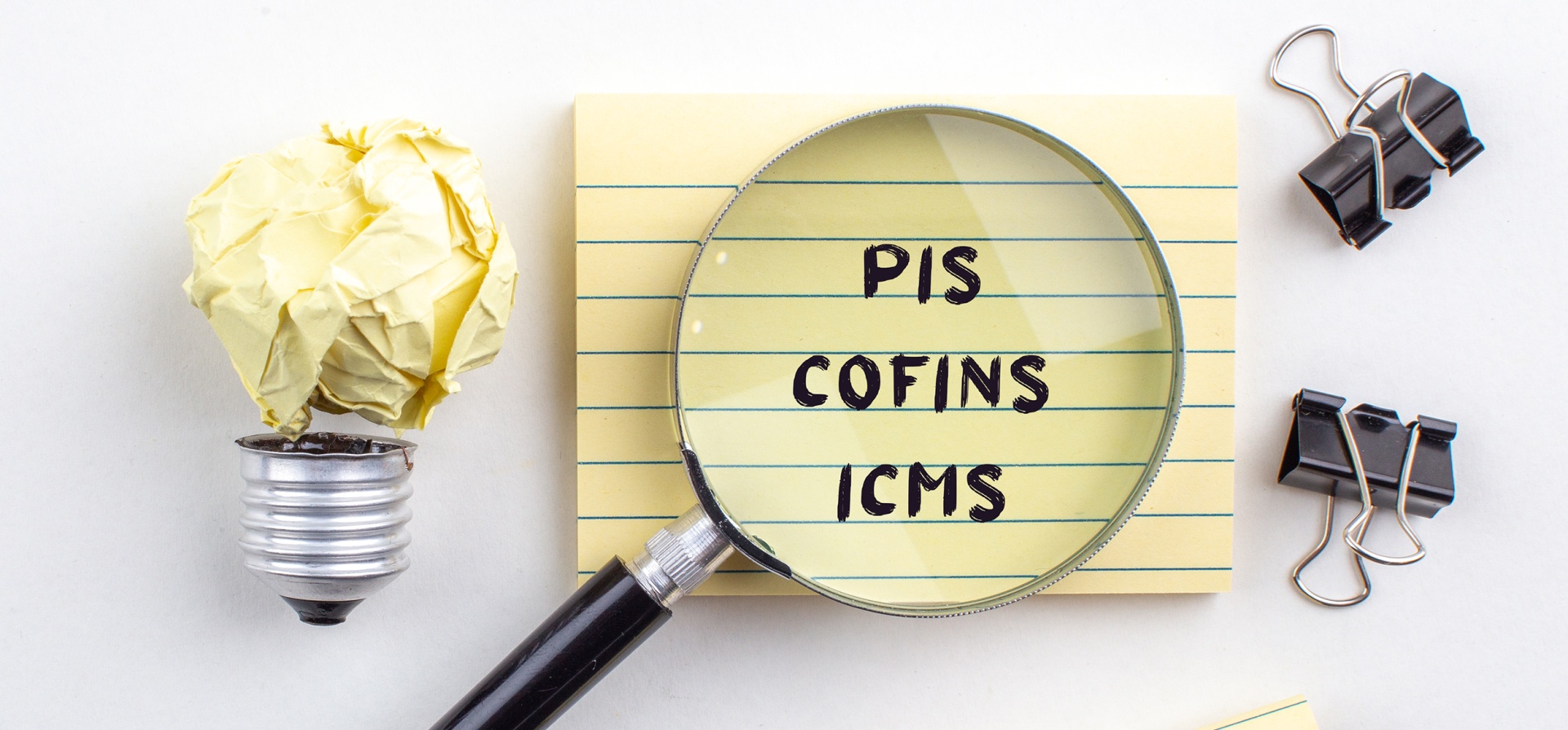 Lupa sobre bloco de notas com os dizeres PIS, Cofins e ICMS ao lado de uma lâmpada feita de papel amassado e grampos de papel pretos.