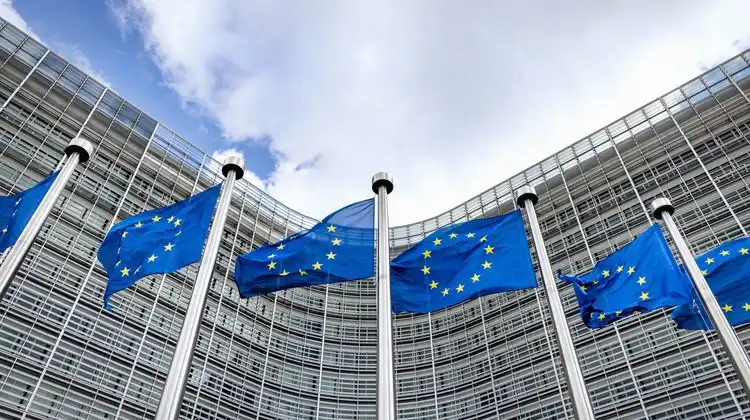 Bandeiras da União Europeia tremulam em mastros em frente à fachada de vidro de um edifício institucional, ilustrando a suspensão de importações de carnes do Brasil.