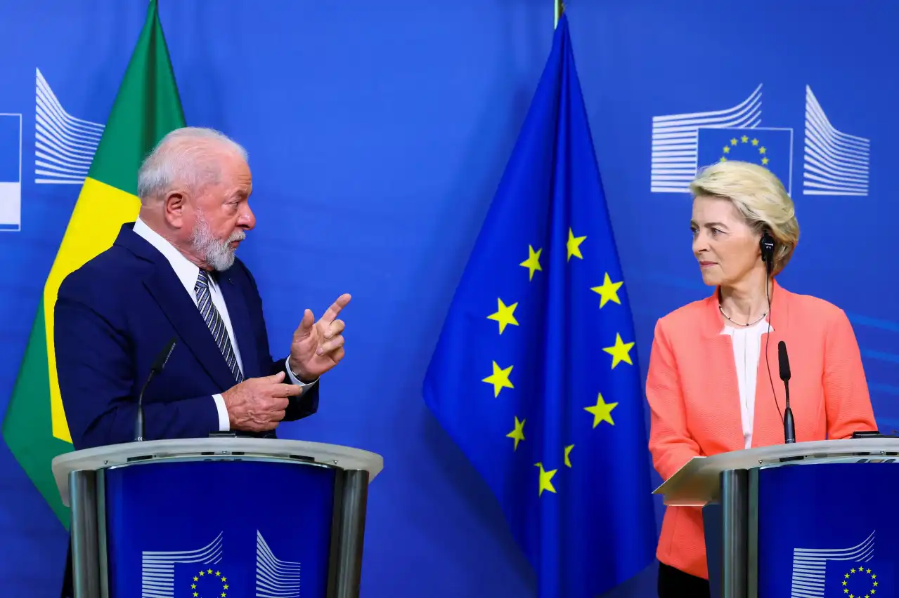 O presidente Lula conversa com Ursula von der Leyen, presidente da Comissão Europeia, em evento oficial com as bandeiras do Brasil e da União Europea ao fundo.