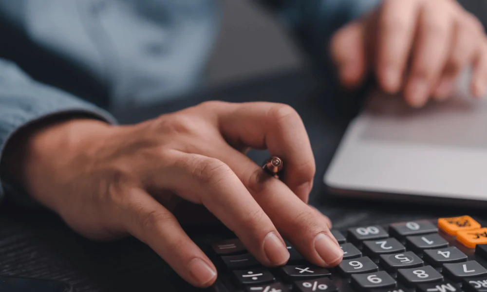 Pessoa calculando números em uma calculadora e usando notebook para planejamento financeiro e análise do orçamento empresarial.