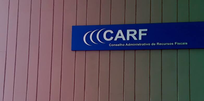 Placa azul com a logomarca do CARF e a inscrição Conselho Administrativo de Recursos Fiscais fixada em uma parede com painéis verticais.