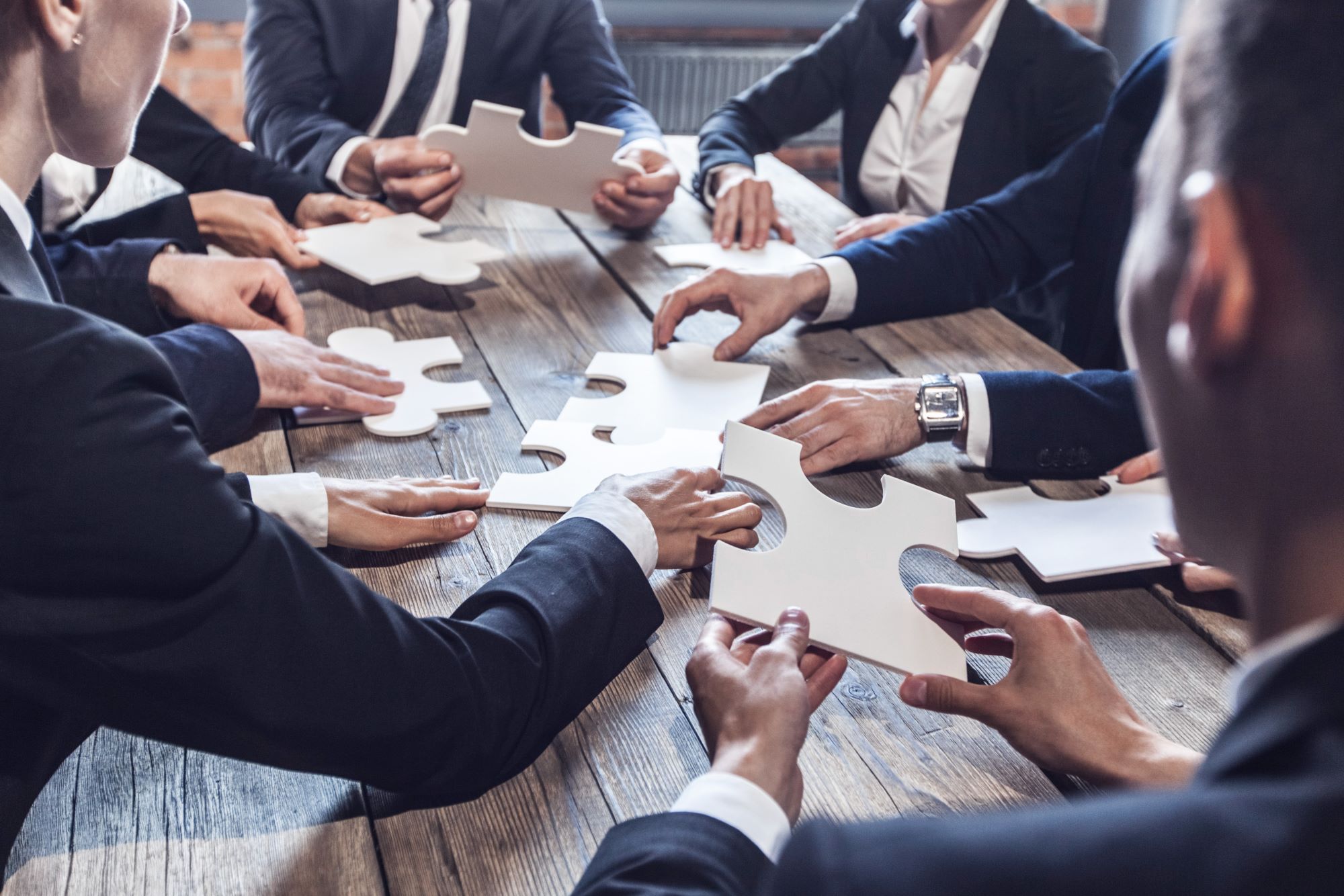 Equipe de empresários de terno reunida ao redor de uma mesa de madeira montando um quebra-cabeça, ilustrando a reorganização da estratégia de negócios frente à Reforma Tributária.