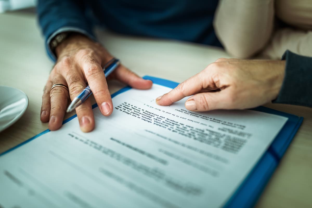 Plano detalhado das mãos de duas pessoas sobre um documento impresso em uma prancheta azul. Uma das pessoas segura uma caneta prateada apontando para as linhas do contrato, enquanto a outra indica um ponto específico do texto com o dedo.