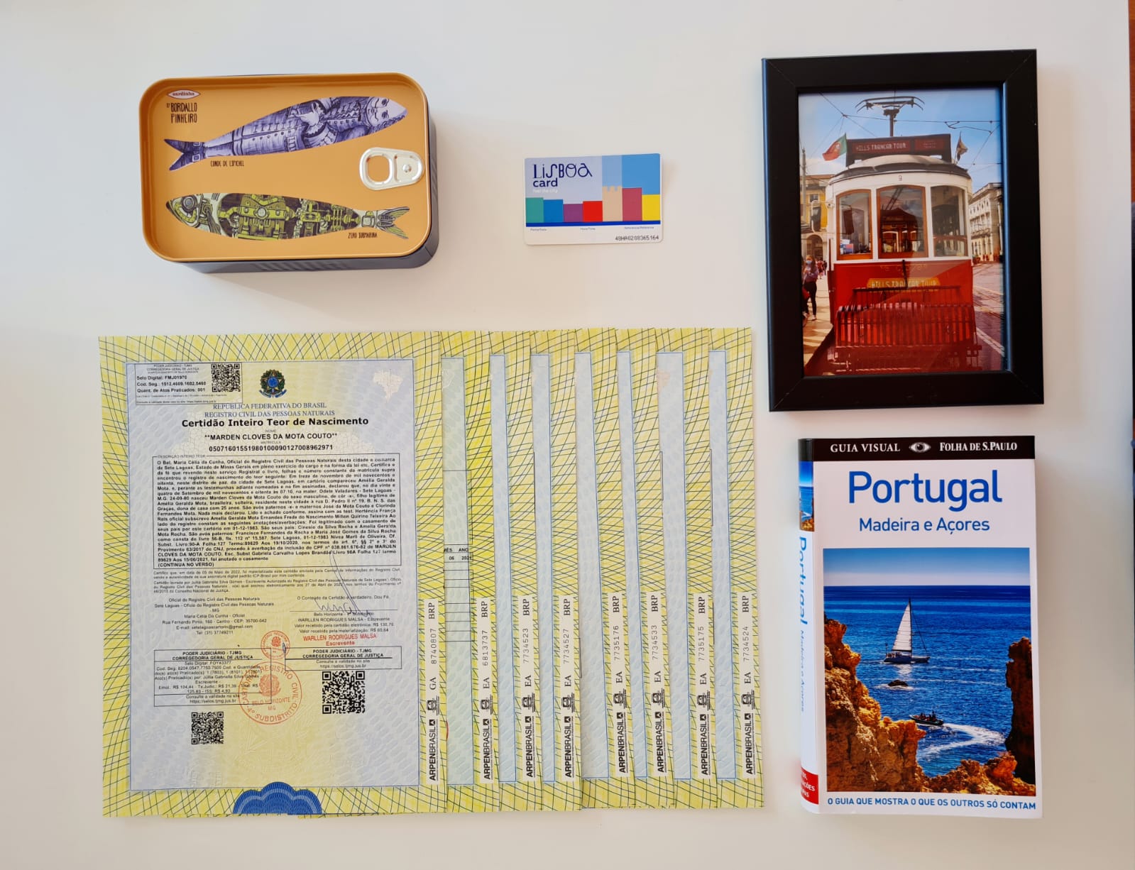 Vista superior de uma mesa com uma Certidão de Inteiro Teor de Nascimento brasileira rodeada por itens de viagem a Portugal, incluindo um guia turístico "Portugal - Madeira e Açores", um porta-retratos com a foto de um bonde elétrico de Lisboa, um cartão "Lisboa Card" e uma lata decorativa.