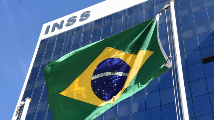 Bandeira do Brasil hasteada em frente ao edifício com a fachada do INSS, ilustrando o período de graça e a manutenção dos direitos previdenciários.