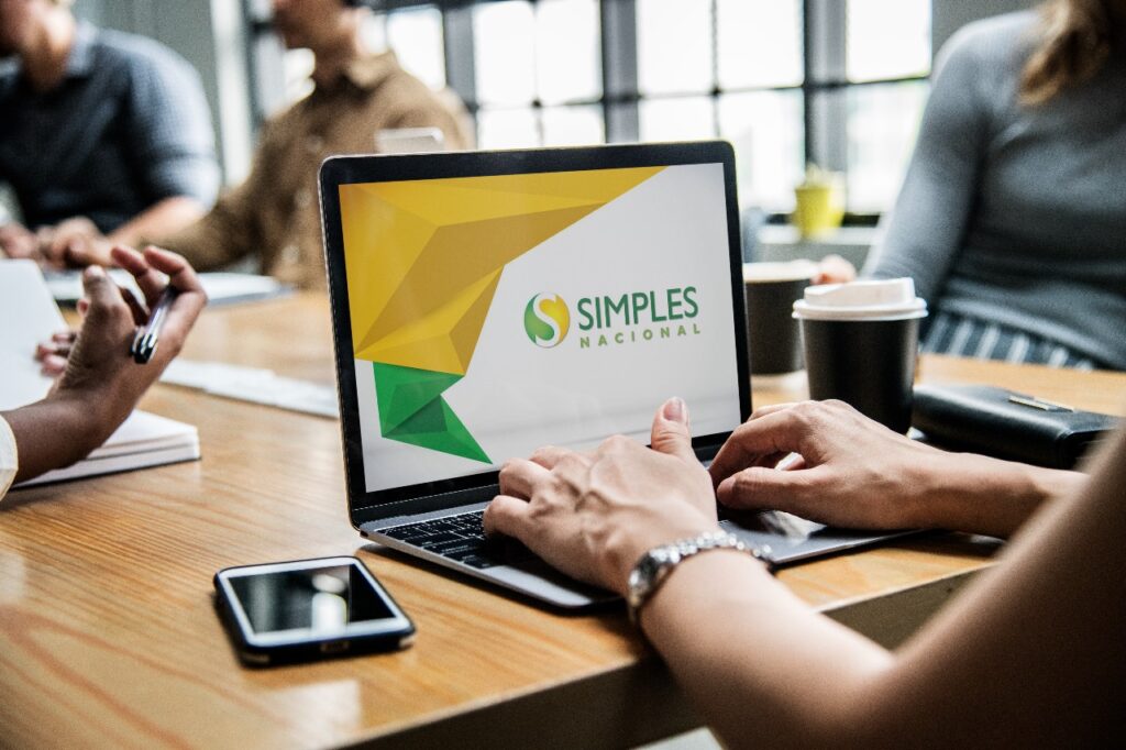 Pessoa digitando em um notebook que exibe a logo do Simples Nacional na tela, em uma mesa de reunião com equipe ao fundo, ilustrando discussões sobre o Simples Nacional 2027 e os impactos da reforma tributária.