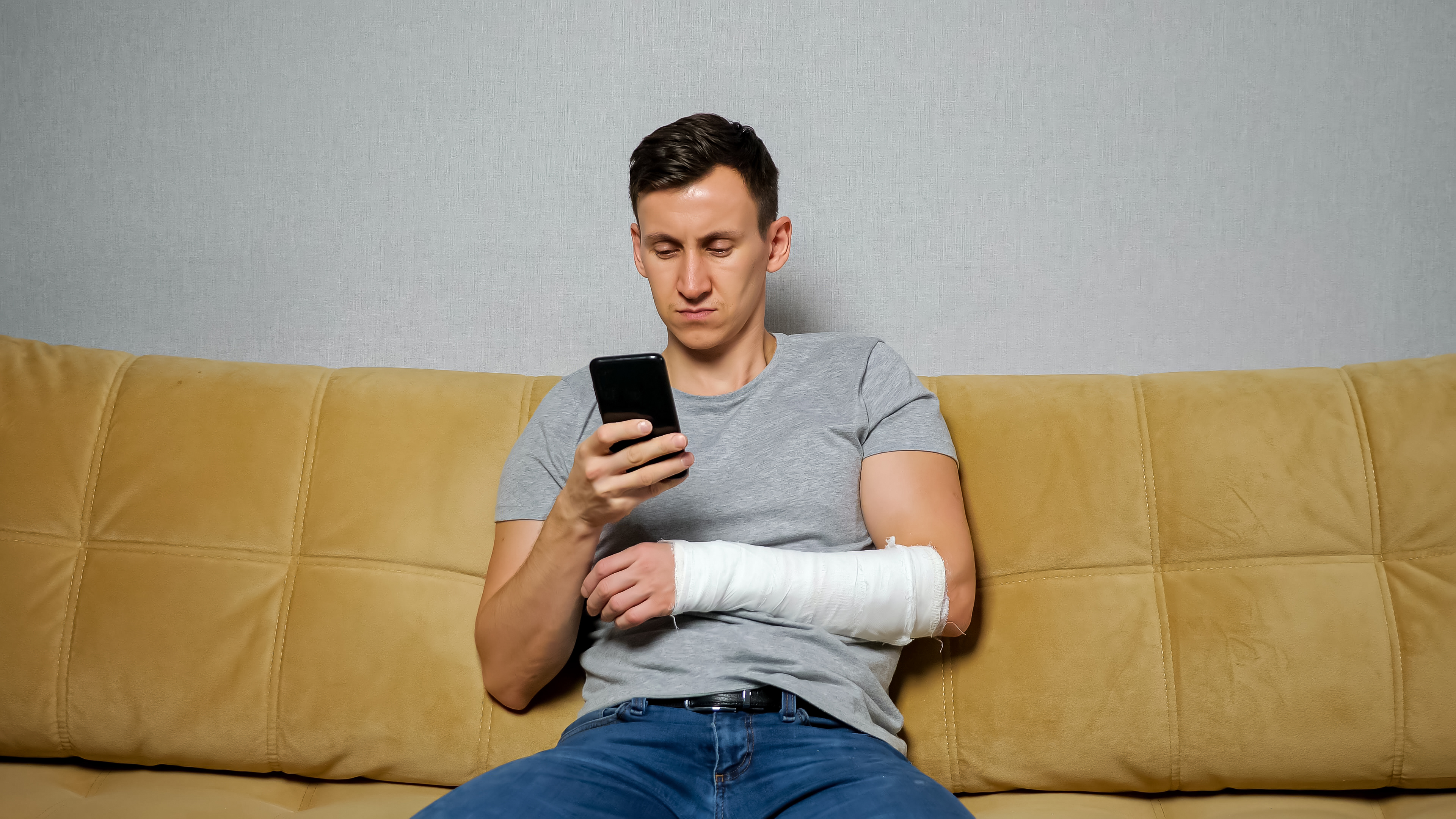 Homem sentado no sofá com o braço engessado mexendo no celular, ilustrando a espera por auxílio do INSS.