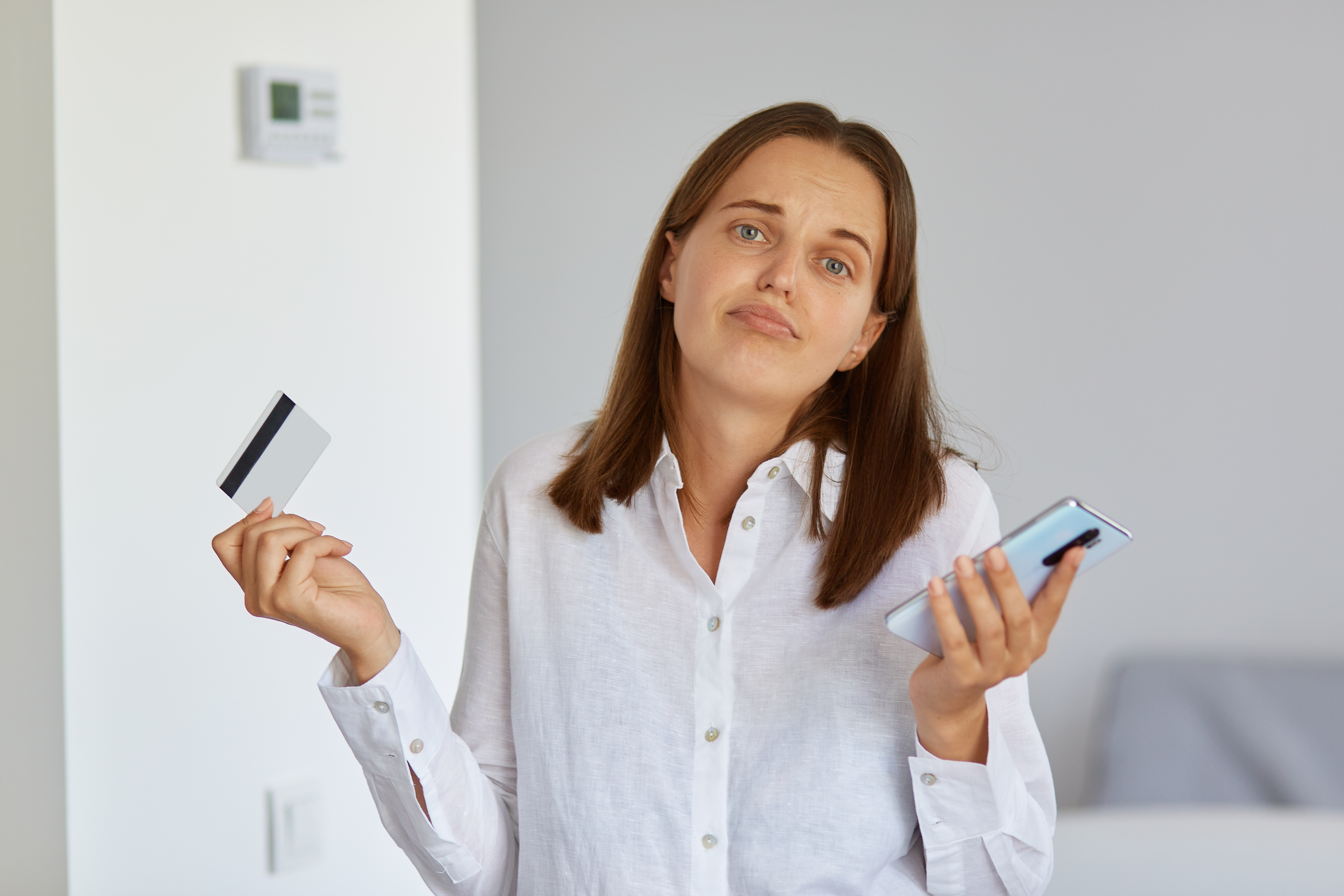 Mulher de branco confusa segura um cartão de crédito e um celular, ilustrando dúvidas sobre custos adicionais como o RAD na retenção de impostos.