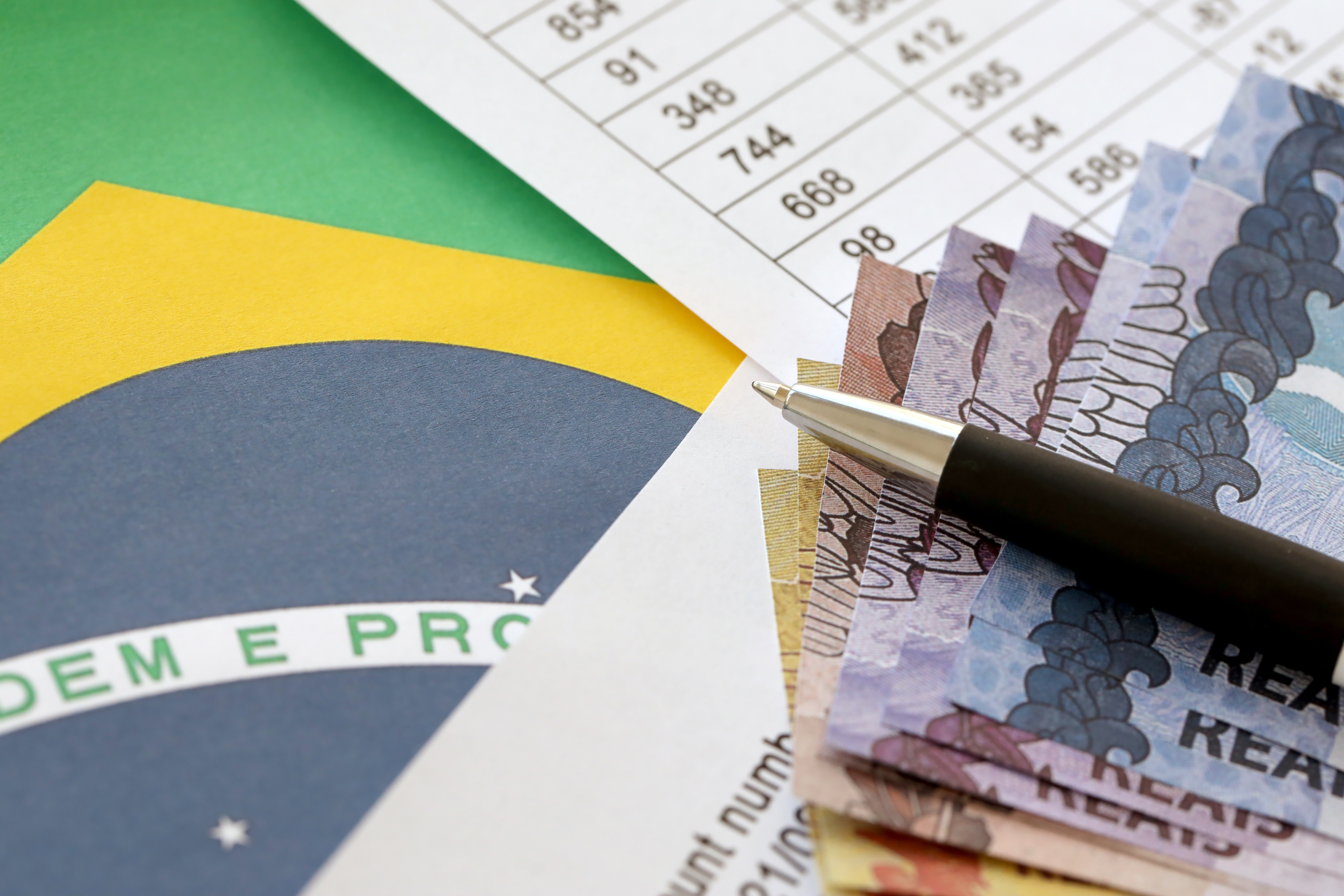 Planilha com números, notas de Real, uma caneta e a bandeira do Brasil ilustram os impactos do imposto seletivo nos custos da empresa.