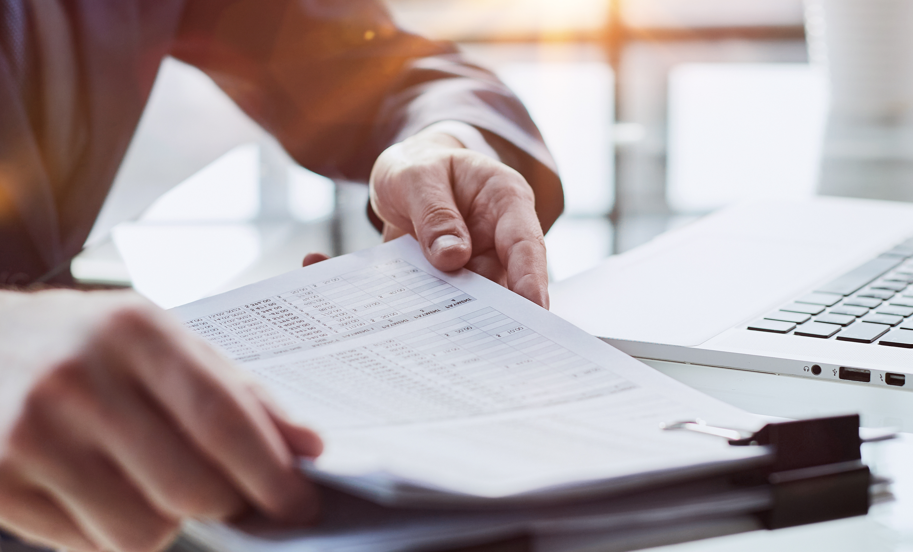 Profissional analisa documentos e notas fiscais de serviços em mesa de escritório ao lado de um notebook.